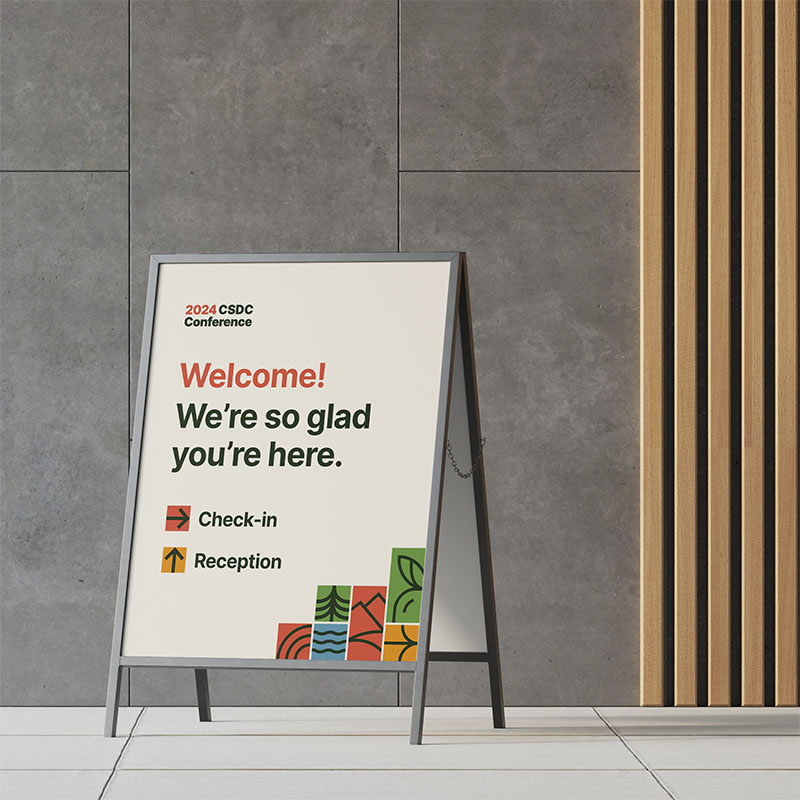 A poster in a stand at check-in, reading Welcome! We're so glad you're here. It's set up on a gray stone floor, with a cement and wood wall behind.