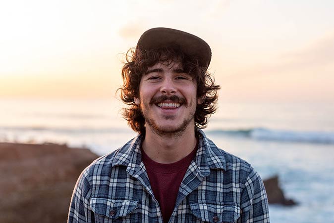 Close-up portrait of Cody wearing a hat and smiling against a blurry background of the coast at sunset